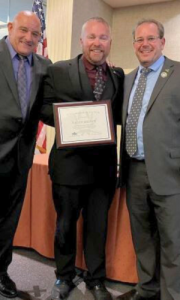 three men in suits standing side-by-side with Geoff Brown in the middle holding a certificate