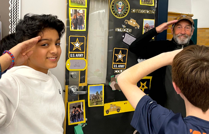 Two students salute a veteran next to a door decorated to represent the United States Army